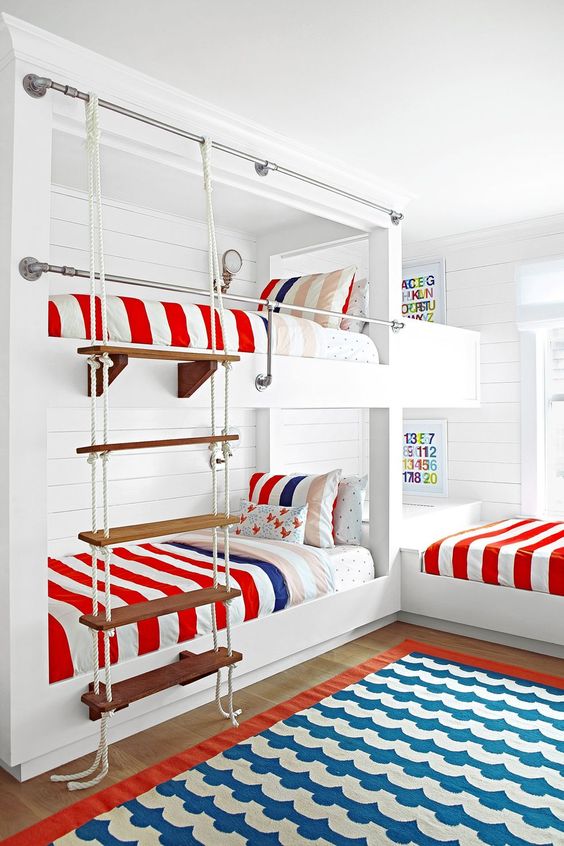 a bright seaside kids' room with multiple bunk beds, bright bedding and a bold rug, a rope ladder and some colorful decor