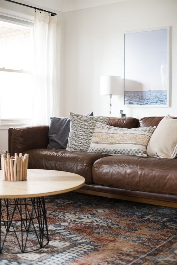 a catchy living room with a brown leather sofa and printed pillows and a rug, a round coffee table and a sea-inspired artwork