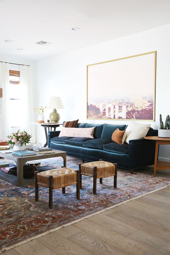 a catchy living room with a navy velvet sofa, a tiered coffee table, woven stools, an oversized artwork and some lamps