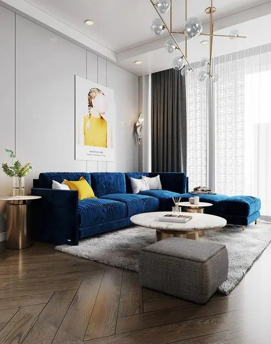 a chic and beautiful living room with dove grey walls, a navy sofa, round tables, a grey pouf, a lovely gold chandelier and some touches of yellow