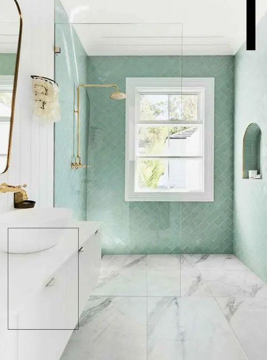 a chic bathroom clad with mint herringbone tiles and marbles ones, a white floating vanity, gold fixtures and natural light