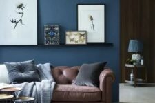 a chic living room with a brown sofa and a navy accent wall plus a ledge gallery wall and an arrangement of side tables