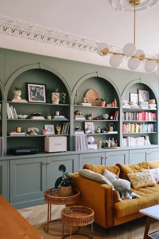 a chic living room with a sage green accent wall with built-in niches and storage units, a mustard sofa, rattan side tables and lovely colorful niche decor