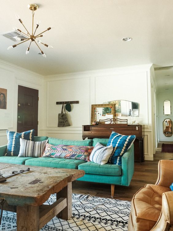 a chic living room with a turquoise sofa and colorful pillows, a vintage piano, a wabi sabi table and a mid century modern chandelier