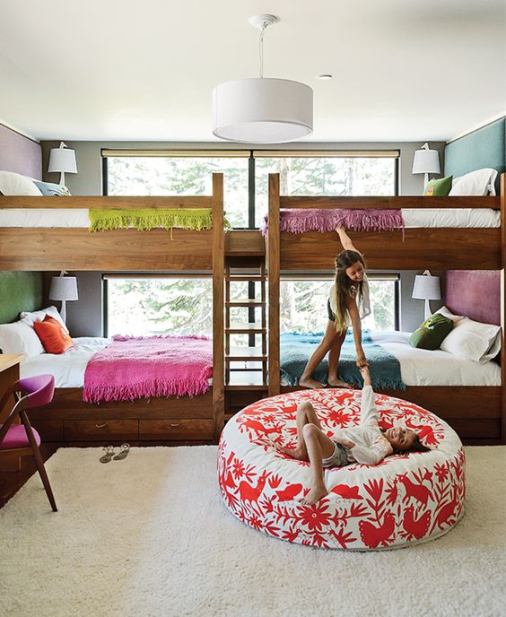 a colorful kids' room with a grey and a purple wall, stained bunk beds, colorful bedding, a round printed pouf and a pink chair