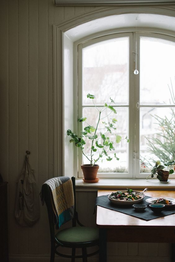 a cozy Scandi dining space with an arched casement window, a stained table and a green chair and potted plants