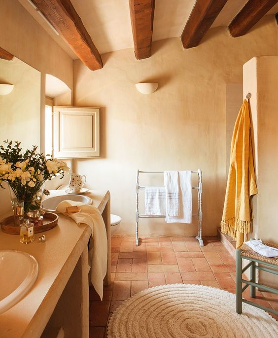a cozy rustic bathroom with plaster walls and terracotta tiles, a large double vanity, a woven stool and wooden beams on the ceiling