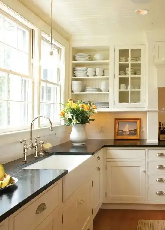 a creamy vintage kitchen with shaker and glass cabinets, black quartz countertops, a window as a backsplash and knobs and handles