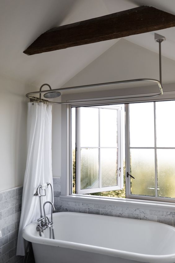 a farmhouse bathroom with grey marble tiles, an oval tub, a casement window and a dark stained wooden beam