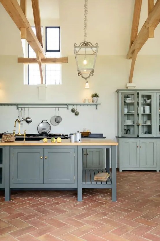 a farmhouse kitchen with a terracotta tile floor, grey cabinetry, a graphite grey kitchen island, wooden beams and pendant lamps