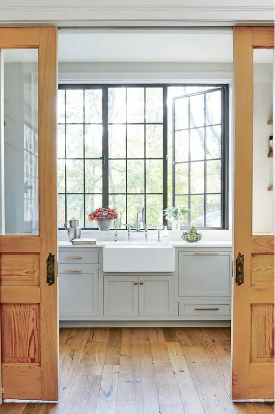 a farmhouse kitchen with grey shaker cabinets, white countertops, a large black frame casement window that lets a lot of light in