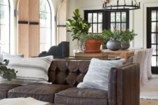 a farmhouse living room with a brown leather sofa, a glass coffee table, a wooden table, black chandeliers and printed pillows