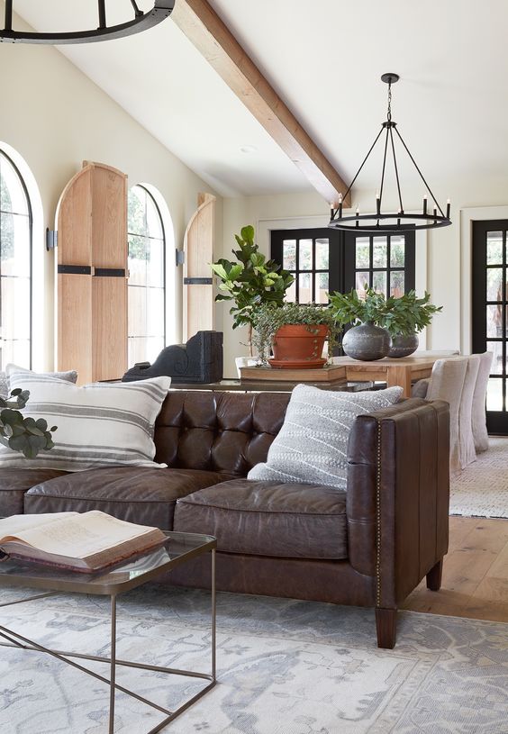 a farmhouse living room with a brown leather sofa, a glass coffee table, a wooden table, black chandeliers and printed pillows