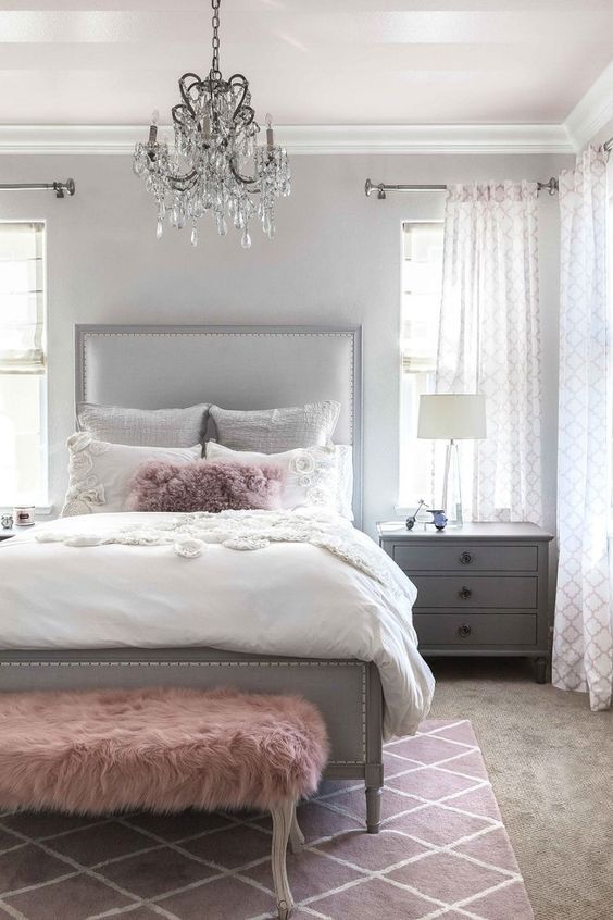 a glam bedroom with a light grey bed and neutral bedding, grey nightstands, a pink faux fur bench and a crystal chandelier