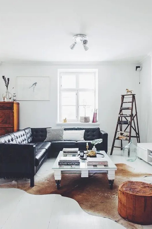 a light filled Scandinavian living room with a black leather sectional, a low industrial coffee table, a white TV unit, a ladder as a storage piece, a rust pouf