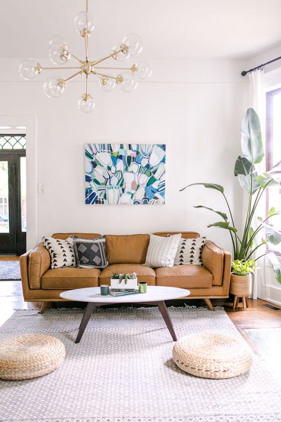 a light-filled living room with a tan leather sofa, a coffee table, jute poufs, a large rug and a bold artwork