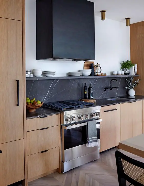 a light stained kitchen with flat panel cabinets, black soapstone countertops and a backsplash, a black hood and black hardware