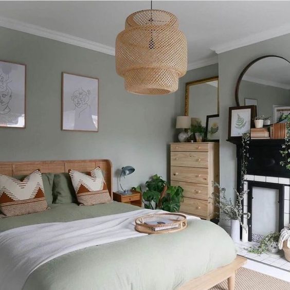 a lovely grey and sage green bedroom with a non-working fireplace, a ratan bed and a stained dresser, potted plants and a woven pendant lamp