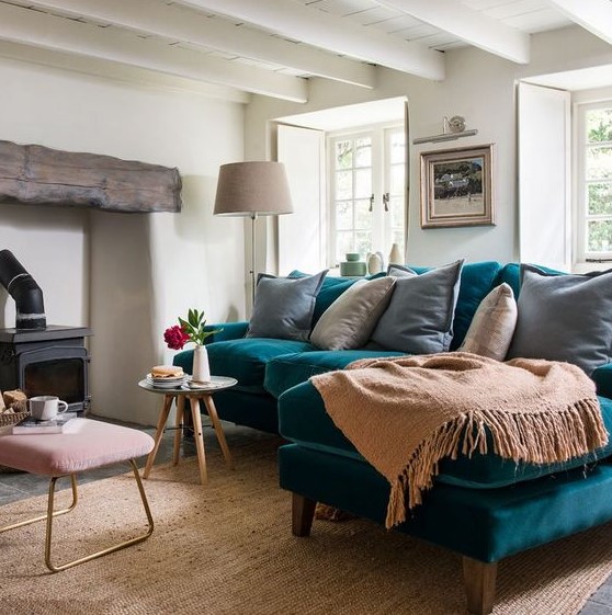 a lovely living room with a hearth with a wooden beam, a teal sofa, neutral textiles, a pink ottoman and a vintage lamp