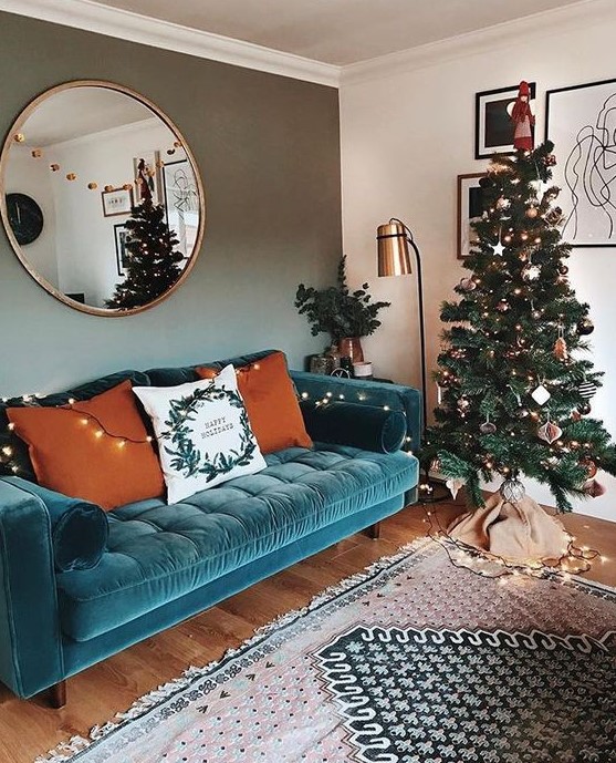 a lovely living room with a turquoise sofa, a Christmas tree with lights, a floor lamp and a round mirror plus a printed rug