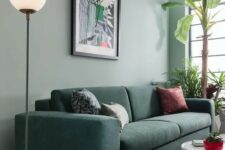 a modern living room with a sage green accent wall, a dark green sofa with printed pillows, a white side table and potted plants