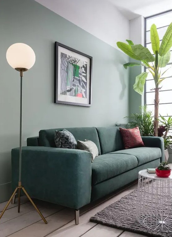a modern living room with a sage green accent wall, a dark green sofa with printed pillows, a white side table and potted plants