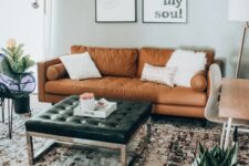 a modern meets boho living room with a tan sofa, a black leather ottoman, a desk and a white chair, a boho rug and potted plants