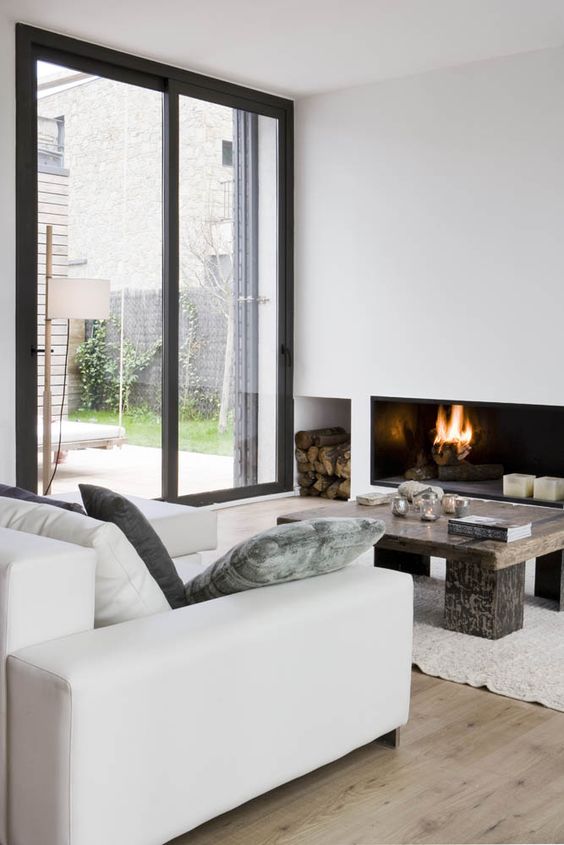 a modern neutral living room with a minimalist fireplace and firewood storage, a wooden table, a creamy sofa and a glazed wall