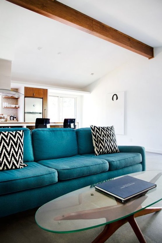 a modern neutral space accented with a rich stained wooden beam, a turquoise sofa with printed pillows, an oval glass coffee table