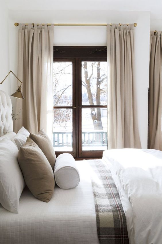 a modern rustic bedroom with a black casement window, a large bed with neutral and printed bedding and a brass sconce