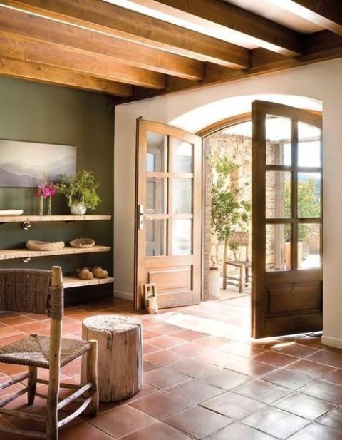 a modern rustic living room with terracotta tiles on the floor, open shelves, a green accent wall, wooden beams and a woven chair