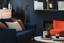 a moody living room with black walls, a faux fireplace, a modern navy loveseat and a bold orange chairs, a round table, a statement artwork and some plants