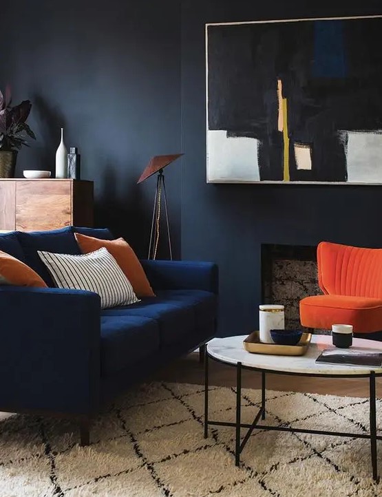 a moody living room with black walls, a faux fireplace, a navy loveseat and a bold orange chairs, a round table, a statement artwork and some plants