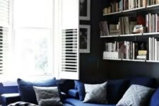 a moody living room with black walls, a navy sectional, printed pillows, open shelves, a leather coffee table, a stylsih mid-century modern chandelier