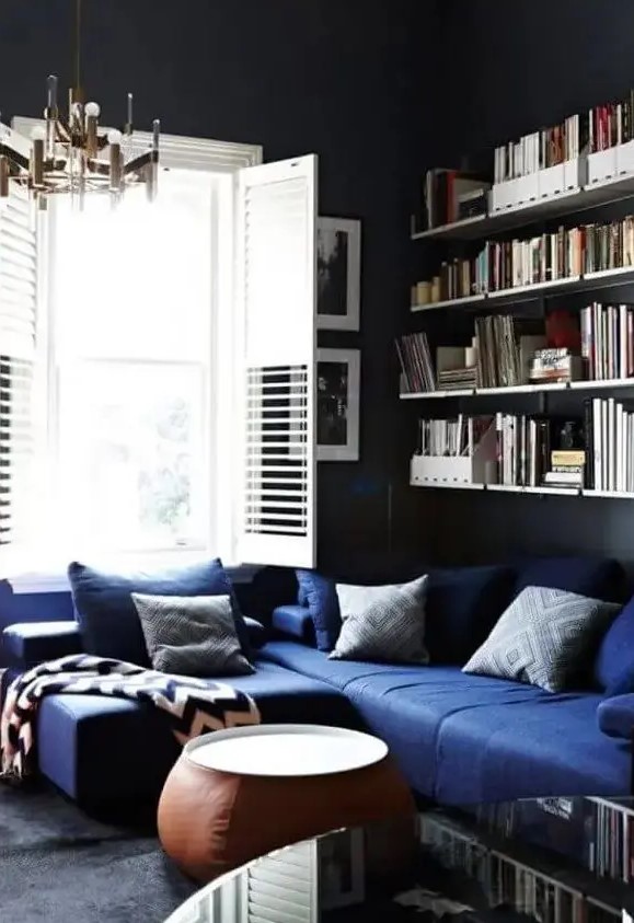 a moody living room with black walls, a navy sectional, printed pillows, open shelves, a leather coffee table, a stylsih mid century modern chandelier