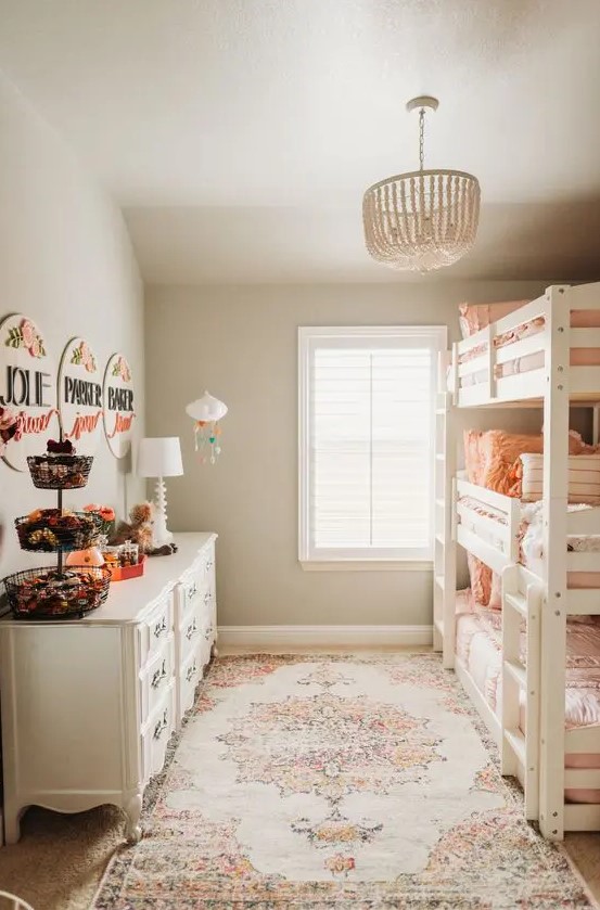 a neutral boho girls' room with a white triple bunk bed with peachy bedding, white dressers, bold and catchy artwork and a bead chandelier