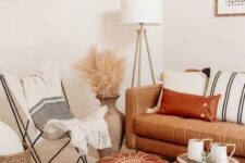 a neutral boho space with a tan leather sofa and a rust-colored ottoman, striped textiles, pampas grass and a wooden table