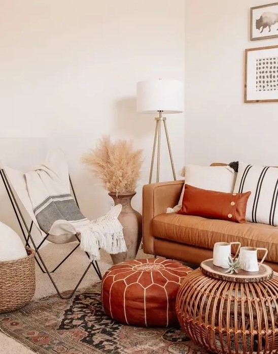 a neutral boho space with a tan leather sofa and a rust colored ottoman, striped textiles, pampas grass and a wooden table