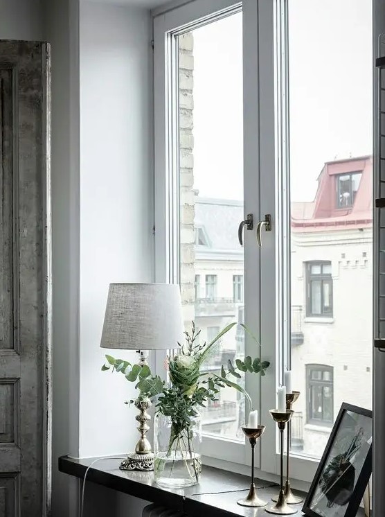 a neutral space with a stylish casement window, a windowsill decorated with candles, a lamp, some greenery and artwork
