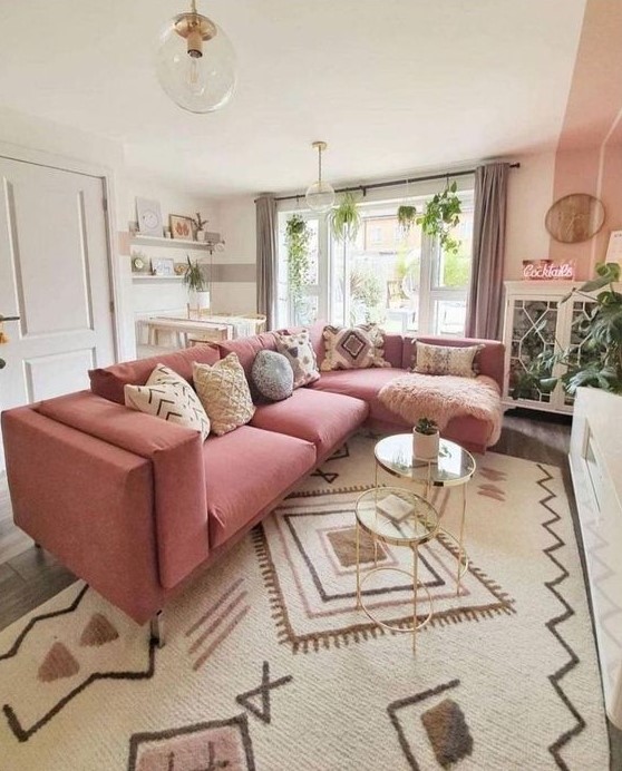 a pretty boho living room done in neutrals, with a large pink sectional in front of the fireplace, glass tables, greenery and a working space by the window