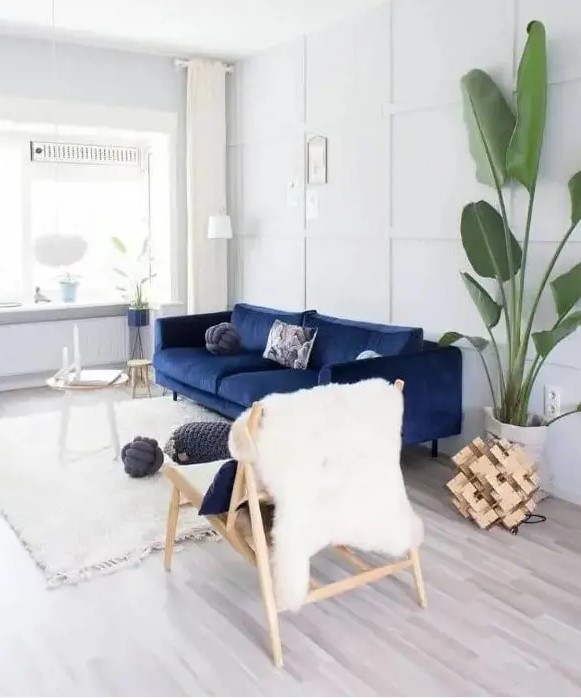 a pretty light-filled lviing room done in white and creamy, with a navy sofa, a couple of side tables, a wooden chair, a potted tree and some wood