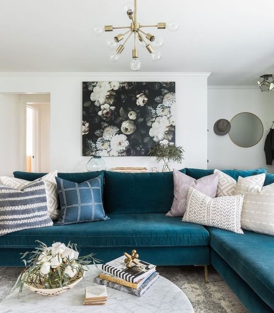 a pretty living room with a teal sectional, a dark floral artwork, a marble coffee table and a gold chandelier
