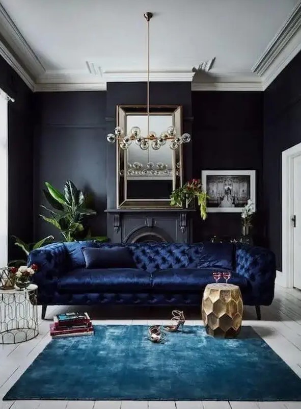 a refined living room with black walls, a non-working fireplace, a navy sofa, a bulb chandelier, a turquoise rug and a gold faceted side table and some books and plants