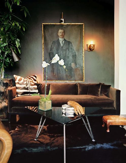 a refined living room with olive green walls, a brown velvet sofa, a triangle coffee table, a leather stool and a potted plant