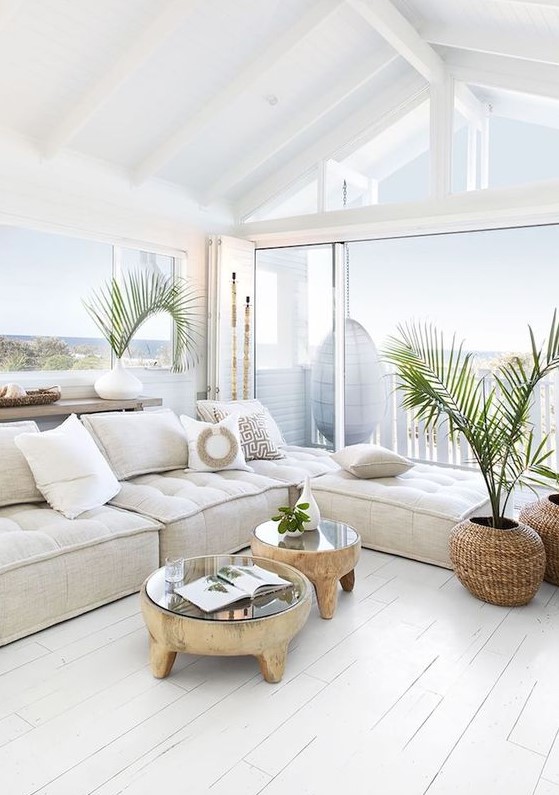 a relaxed coastal living room in white, with a low sectional, wood and glass coffee tables and potted plants plus a sea view