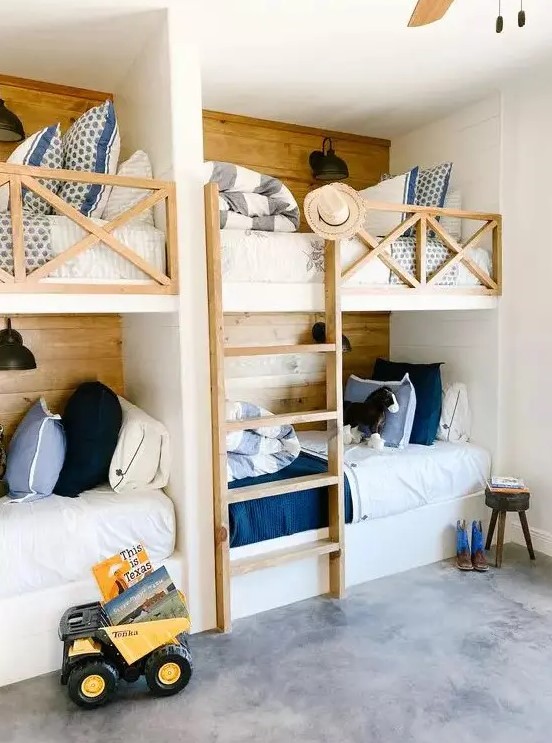 a rustic kids' room with four built in bunk beds, white, blue and navy bedding, a ladder and lots of pillows and black sconces