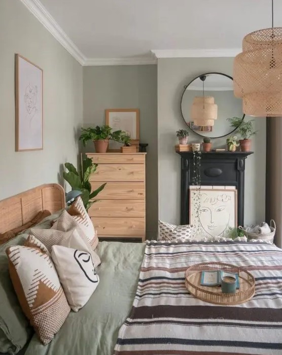 a sage green bedroom with stained furniture, printed bedding, a non-working fireplace, a woven pendant lamp and potted greenery
