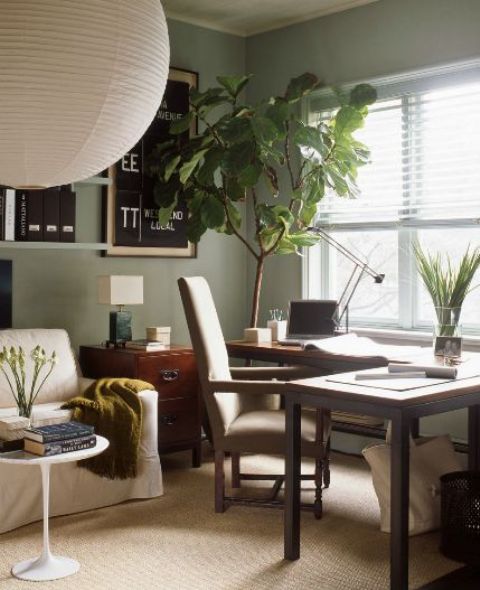 a sage green home office with a corner desk, an upholstered chair, a neutral sofa, potted plants, open shelves and a memo board