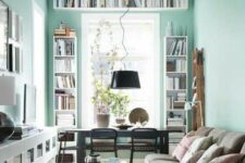 a small living and dining room with mint blue walls, bookshelves, a dining space by the window, a sofa and a floating credenza