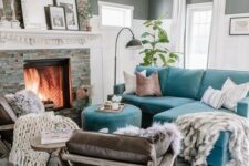 a small living room with grey walls, a fireplace clad with faux stone, a turquoise sofa with a pouf, leather chairs and blankets